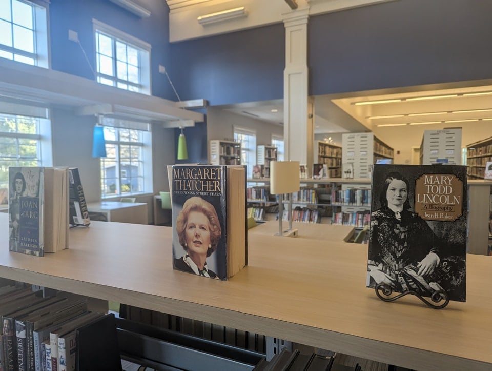 Books about historical women, including Margaret Thatcher and Mary Todd Lincoln, are displayed on wooden shelves in a well-lit library with large windows and fluorescent lights.