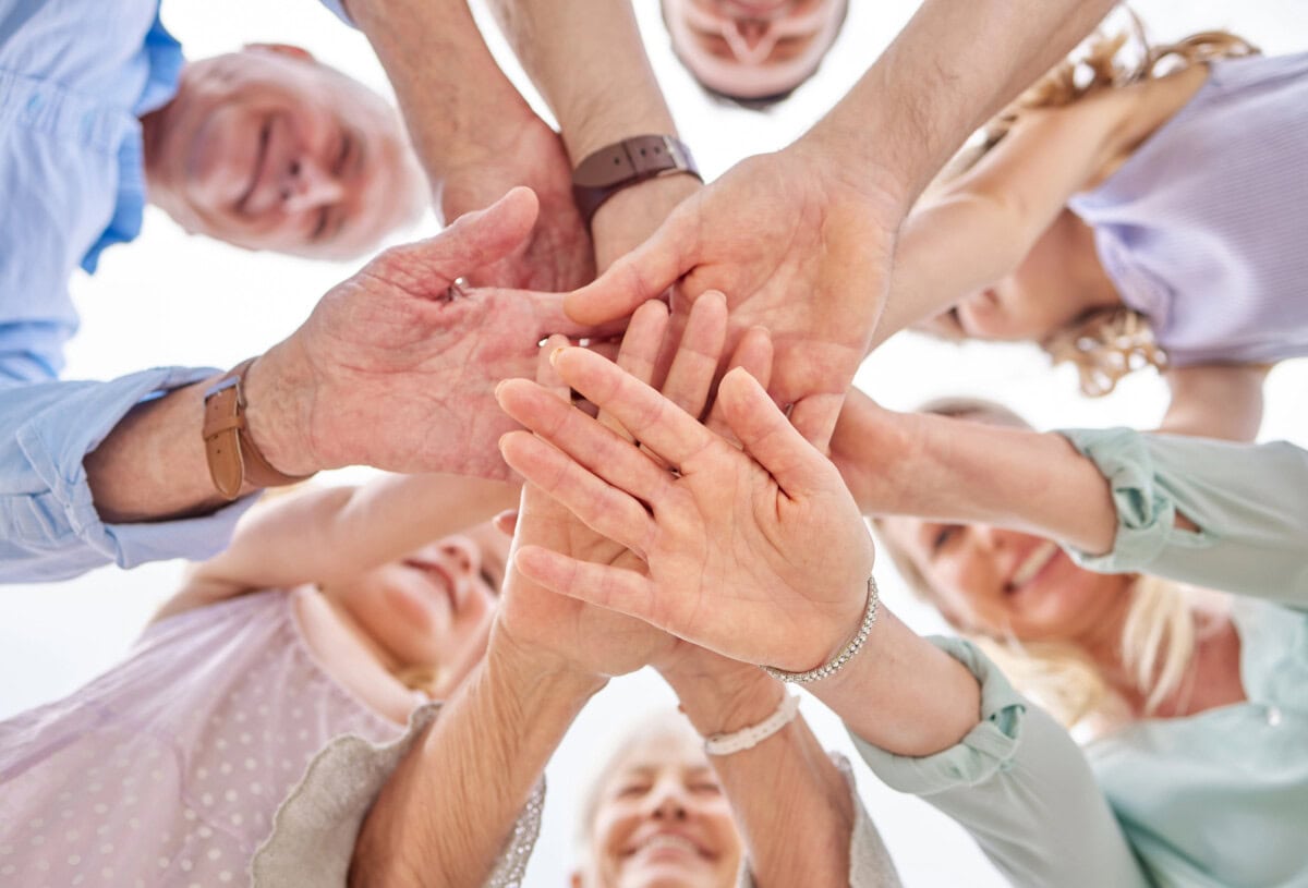 a group of people standing in a circle stack their hands together in the center, viewed from below against a bright sky.