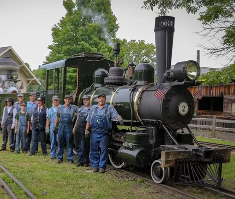 Hesston Steam Museum: A group of people in work clothes stands beside a vintage black steam locomotive on railroad tracks, with trees and buildings in the background.