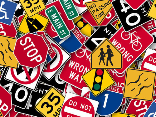 A variety of overlapping road signs including stop, speed limits, street names, wrong way, pedestrian crossing, no parking, and traffic light symbols.