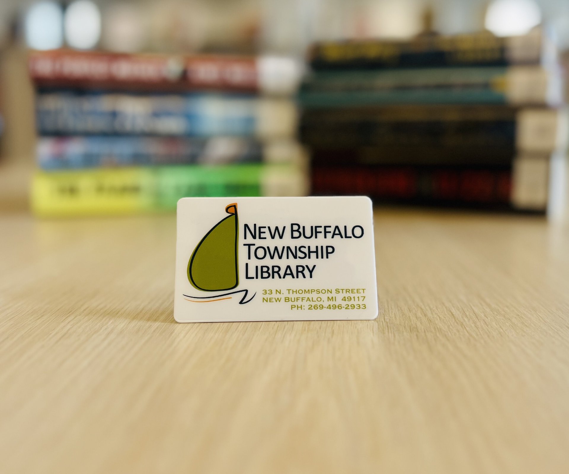 Picture of a New Buffalo Township Library Card on a beige tables, with two stacks of books blurred in the background
