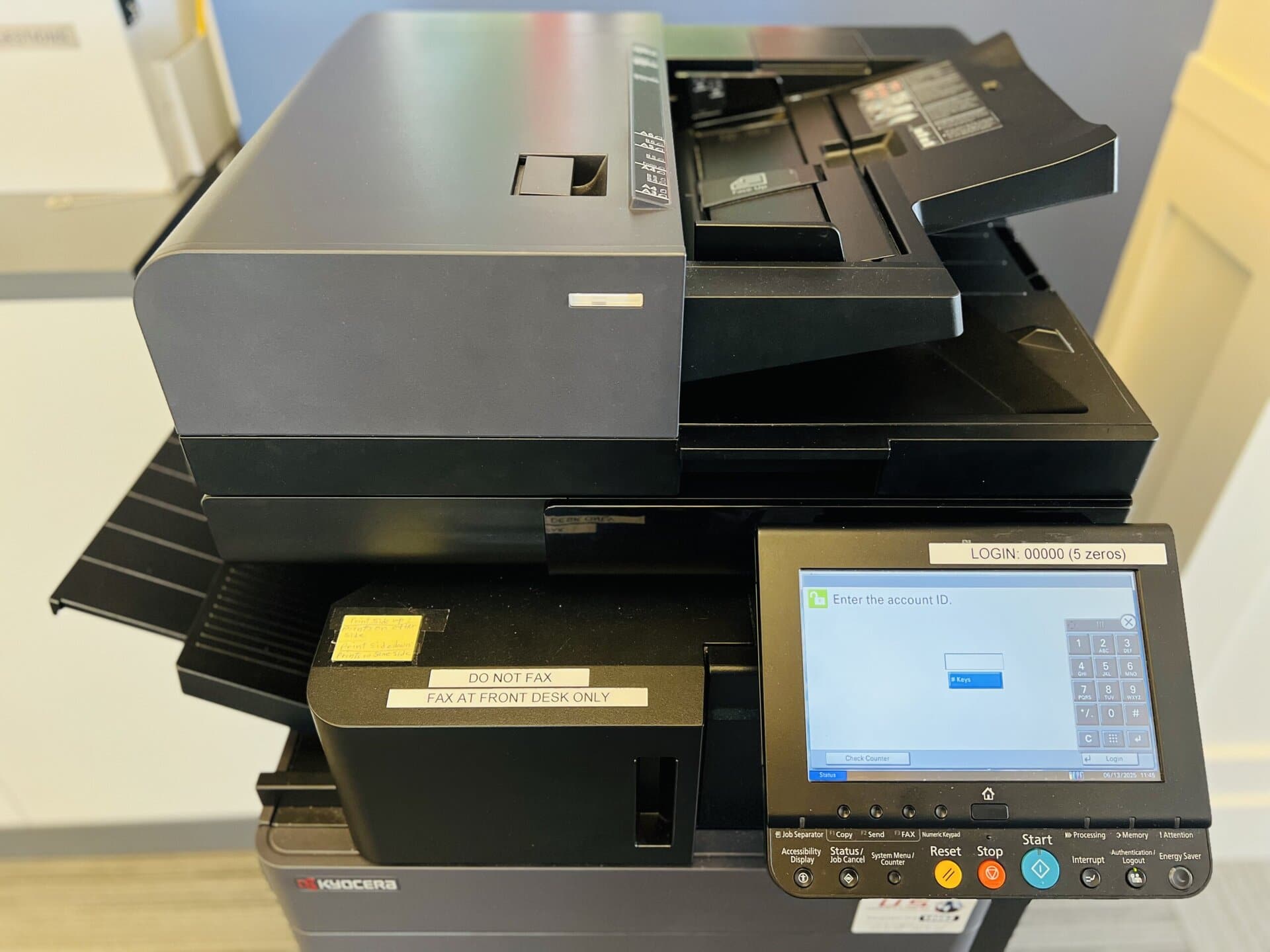 Black Kyocera multi-function printer and copier with a touchscreen login screen and keypad. A label above the screen instructs users to log in with five zeros. Another label on the machine reads "Do not fax; fax at front desk only."