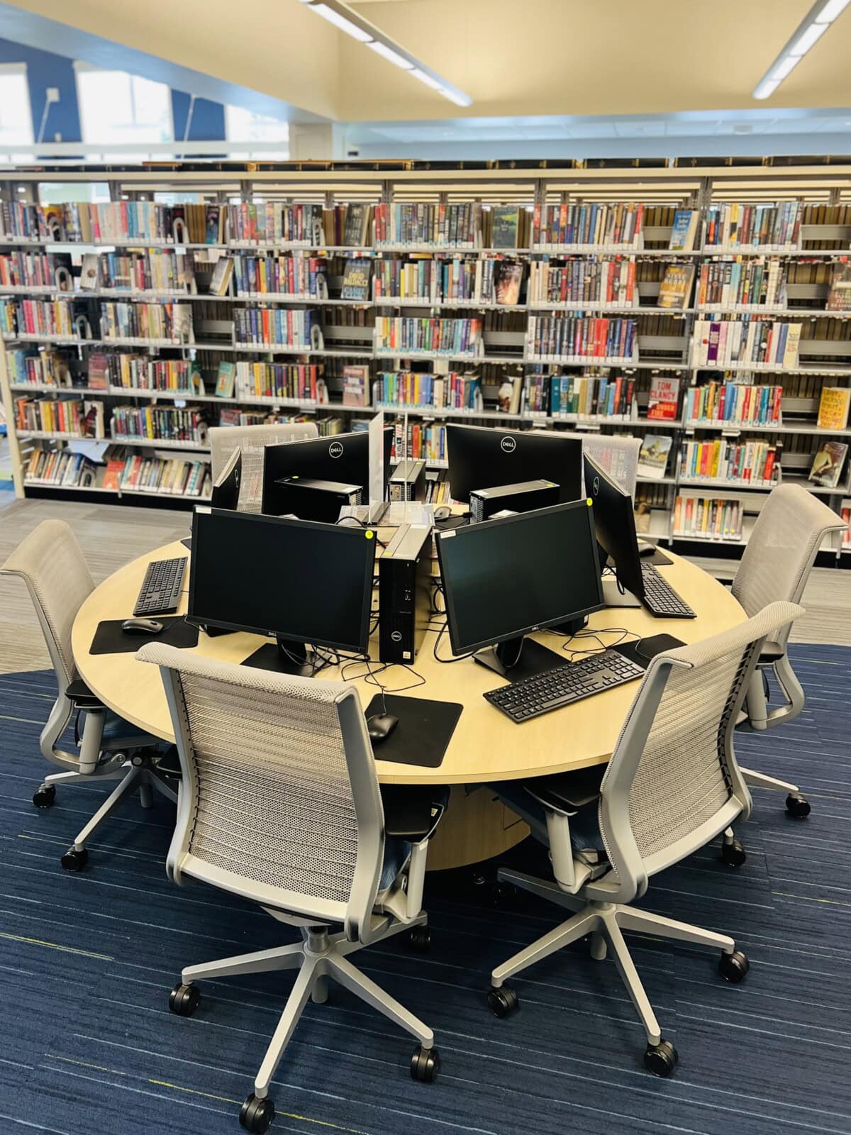 Round table with six computer workstations and office chairs on a blue carpet, located in front of bookshelves filled with colorful books in the library’s main area.