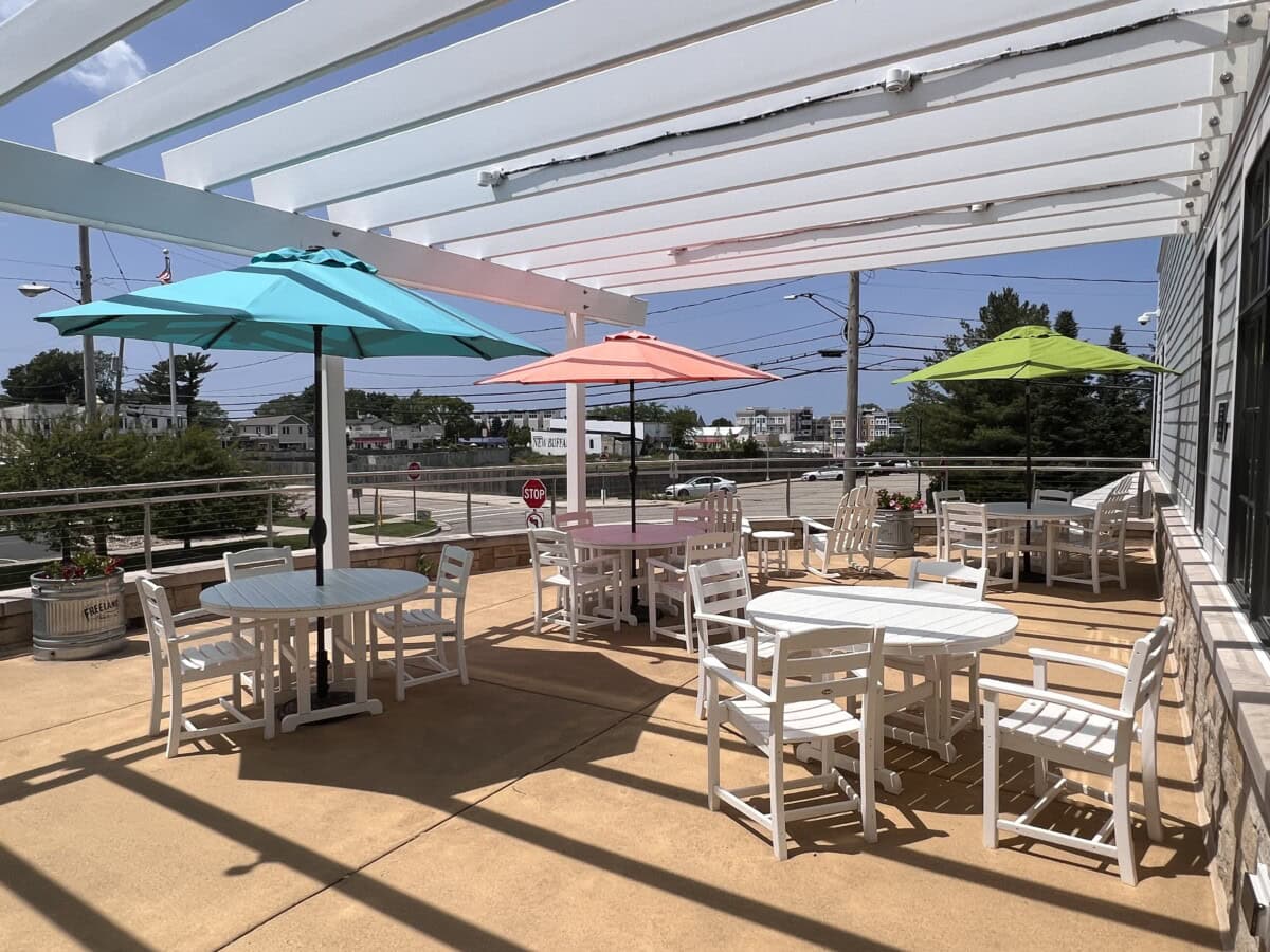 Sunny outdoor patio beneath a white pergola, centered with white tables and chairs arranged evenly under teal, coral, and green umbrellas.