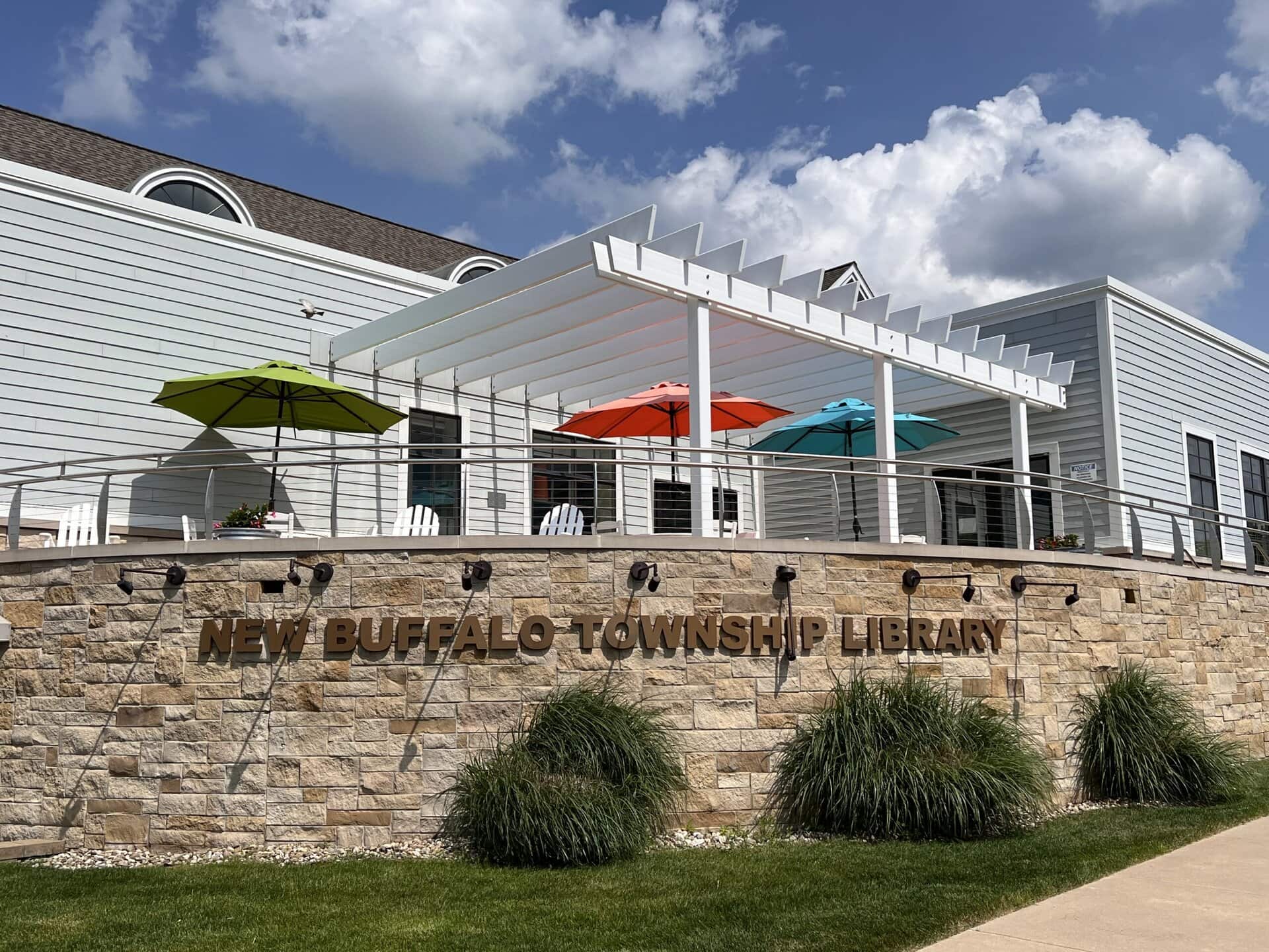 Exterior of the New Buffalo Township Library with a white pergola and colorful patio umbrellas. A stone wall in the foreground features raised metal lettering that reads 'NEW BUFFALO TOWNSHIP LIBRARY.'
