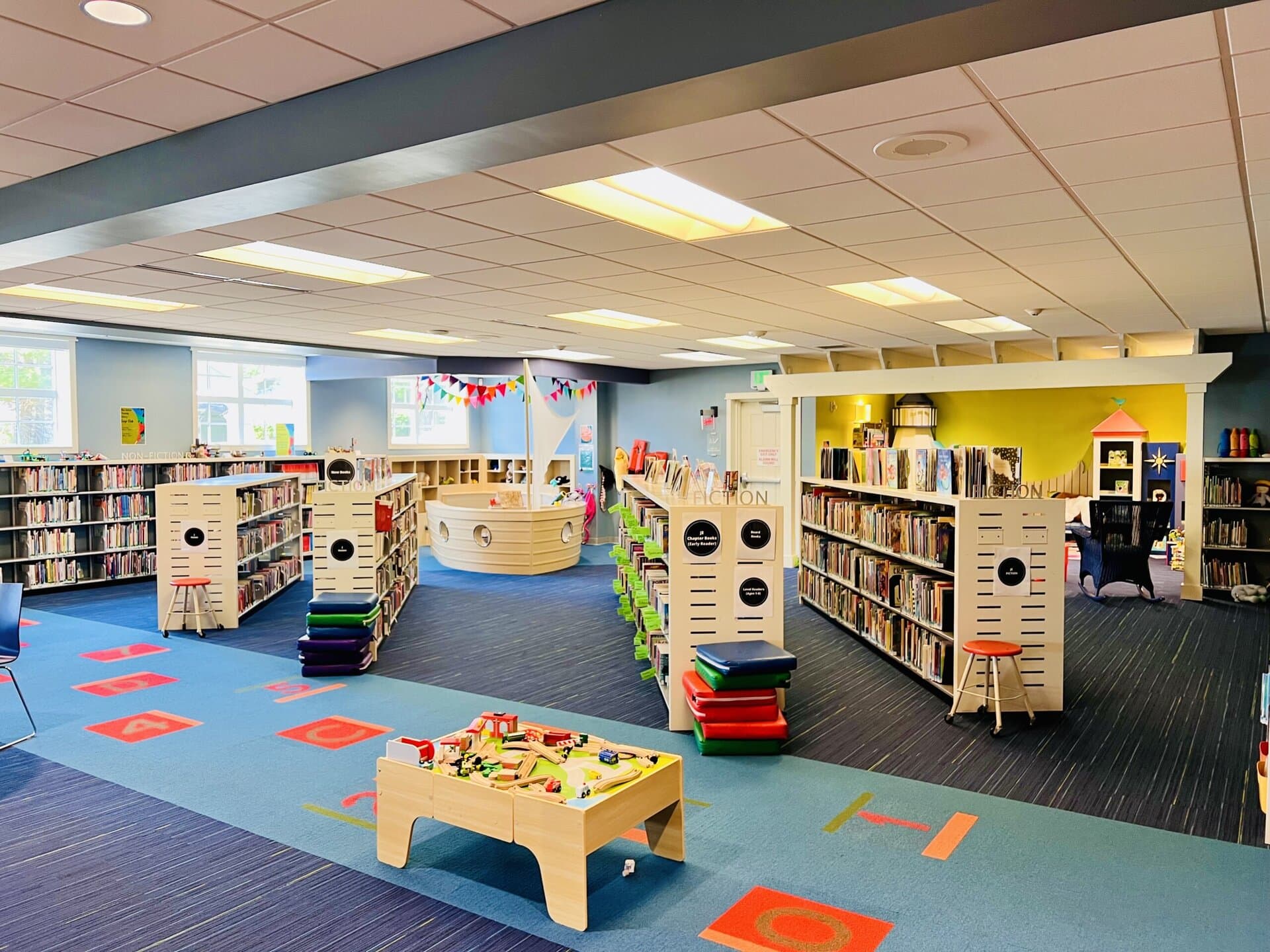 Colorful children’s section of the New Buffalo Township Library, with low bookshelves, reading nooks, a wooden play ship, and a train table centered on a vibrant carpet with hopscotch markings.