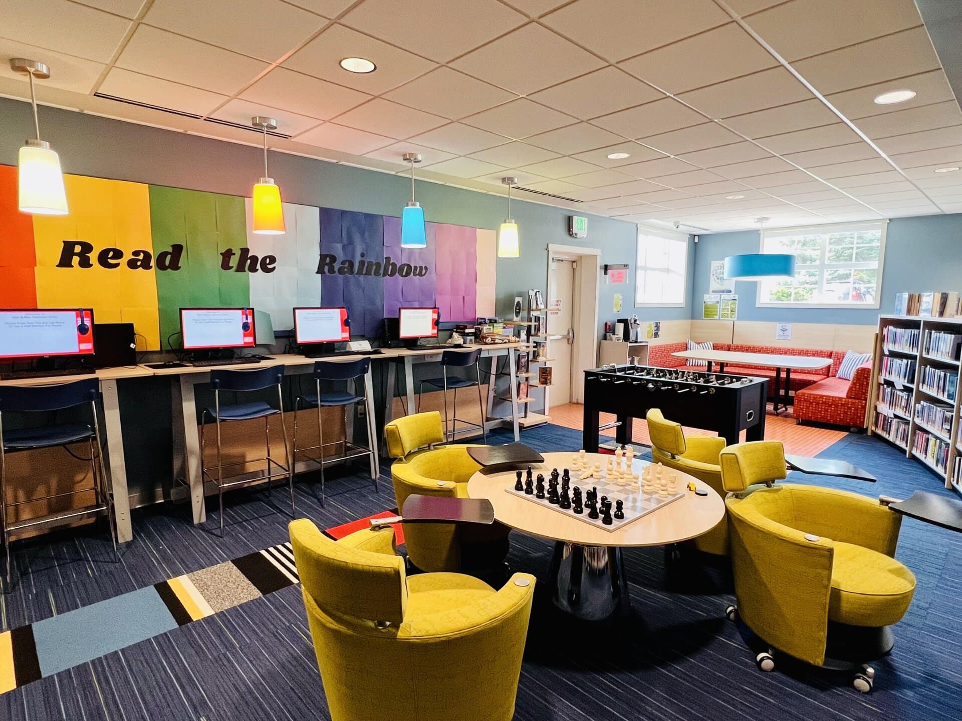 Library teen area with rainbow-colored wall reading 'Read the Rainbow' above computer stations, a central table with a chessboard surrounded by yellow chairs, a foosball table, and a cozy seating nook near the windows.