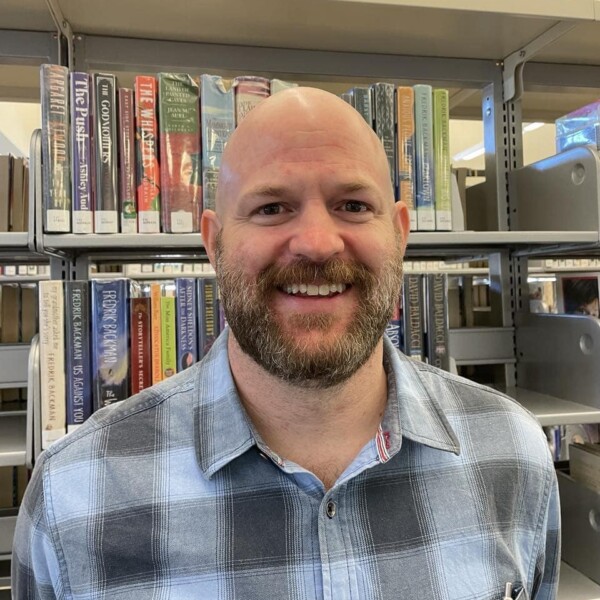 A smiling man with a beard and bald head wearing a plaid shirt stands in front of bookshelves in a library.