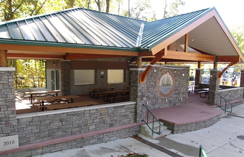 New Buffalo Township Playground: Pavilion with stone walls, green metal roof, picnic tables, and a serving window, located in a wooded park area.