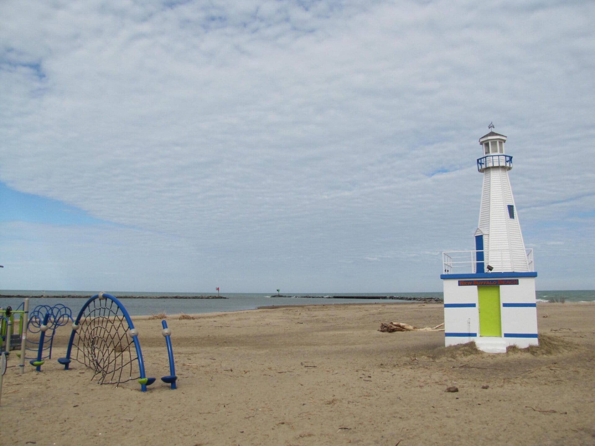 New Buffalo Beach: A sandy beach with a white and green lighthouse structure and a blue playground climbing frame, under a mostly cloudy sky by the water.