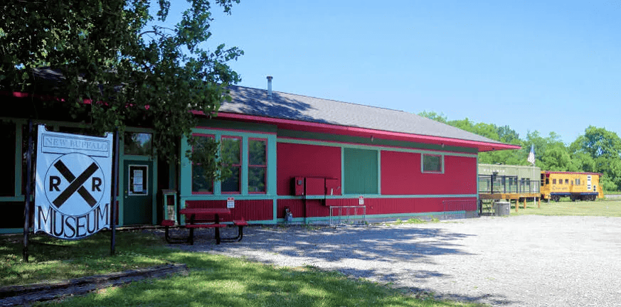 New Buffalo Railroad Museum: A red and green building labeled as a railroad museum sits next to railroad tracks and an old yellow railcar on a clear day.
