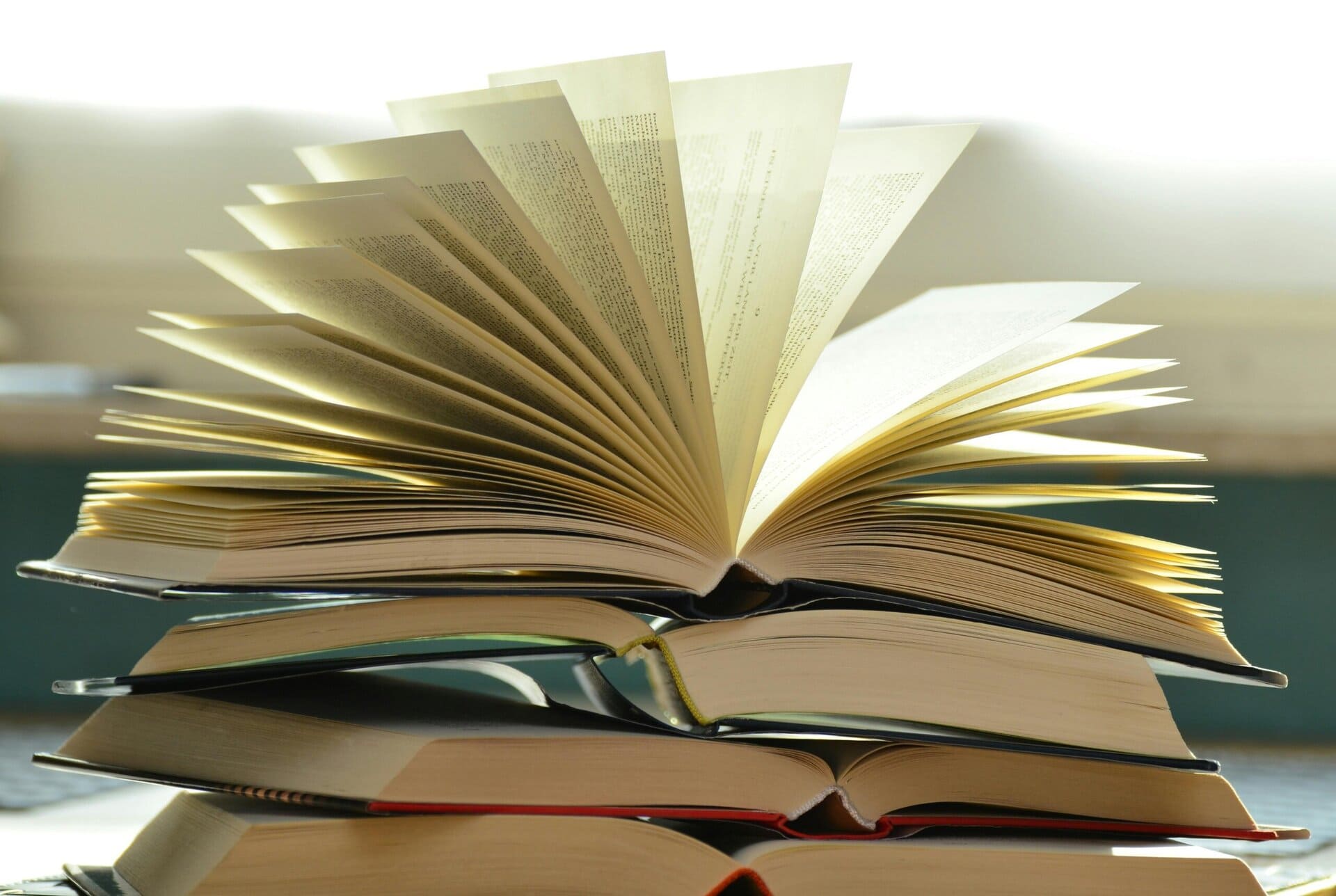 Stack of hardcover books with one book open on top, its pages fanned out in mid-turn, backlit by soft daylight.